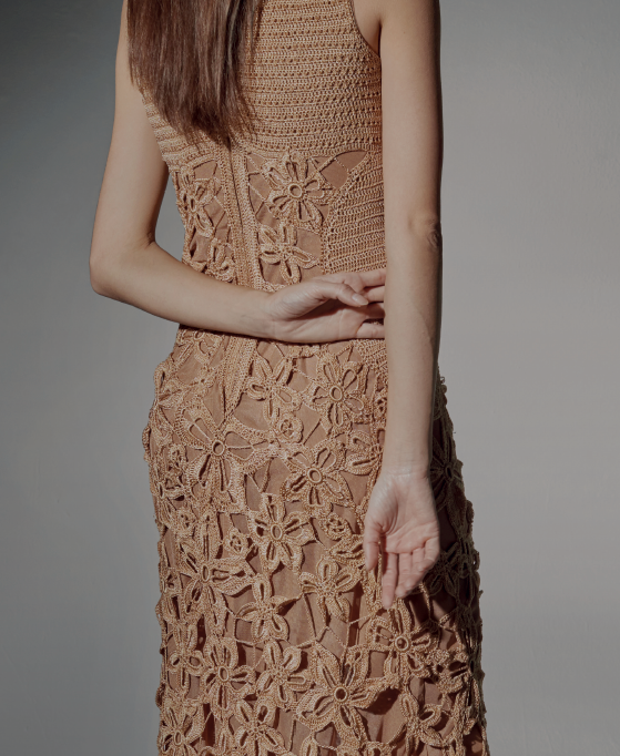 Modelo de frente luciendo el Vestido Ara Berà largo de crochet color caramelo, con escote redondo y flores inspiradas en el toborochi, junto a una pared iluminada por el sol.