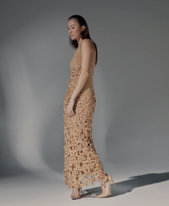 Modelo de frente luciendo el Vestido Ara Berà largo de crochet color caramelo, con escote redondo y flores inspiradas en el toborochi, junto a una pared iluminada por el sol.